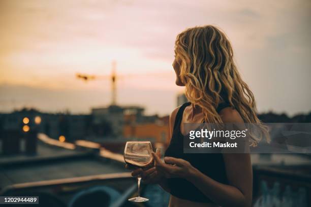 thoughtful young woman having wine while looking away on terrace during rooftop party at sunset - drink stock-fotos und bilder