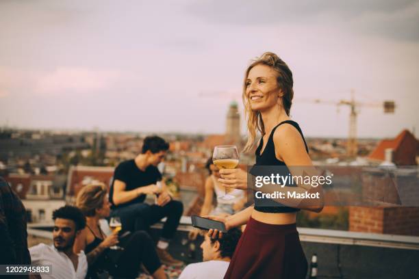 smiling woman enjoying drink while partying with friends at rooftop - dachparty stock-fotos und bilder