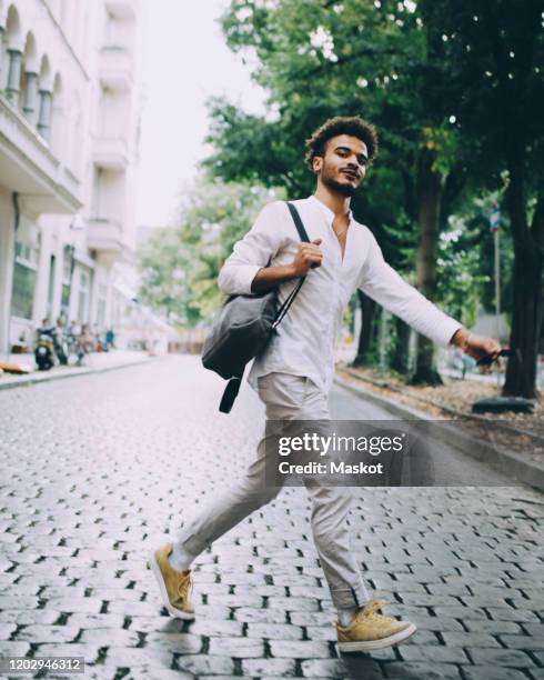 full length of young man carrying backpack while walking on cobbled street in city - man full length stock pictures, royalty-free photos & images