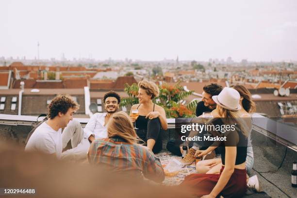 friends enjoying beer while sitting together on terrace at rooftop party - terraza fotografías e imágenes de stock