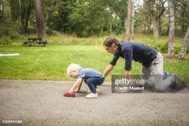 father kneeling while son playing with toy car on footpath against plants at park - toy car stock pictures, royalty-free photos & images
