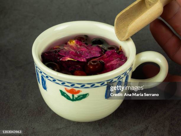 a cup with dish of hot infusion of medicinal herbs with rose hips, hawthorn and rose petals and a hand with a wooden shovel - hot hips 個照片及圖片檔