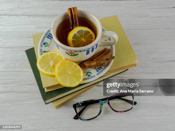 a cup with dish of hot tea with a tea bag, cinnamon and lemon on top of books and reading glasses - herbal tea bag stock pictures, royalty-free photos & images