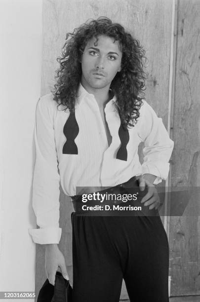 British singer Howcher wearing an open-necked white shirt with an undone bow tie draped around his neck as he leans against a white wall in a studio...