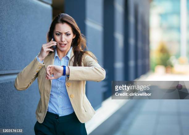 jeune femme d'affaires choquée contrôlant le temps sur la montre-bracelet tout en parlant le téléphone portable - presse photos et images de collection