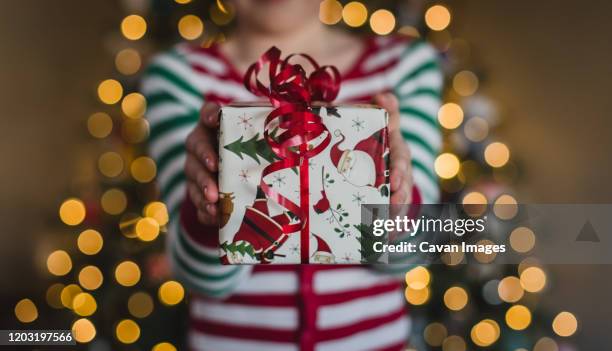 child in pyjamas holding christmas gift in front of lights on tree. - child giving gift stock-fotos und bilder