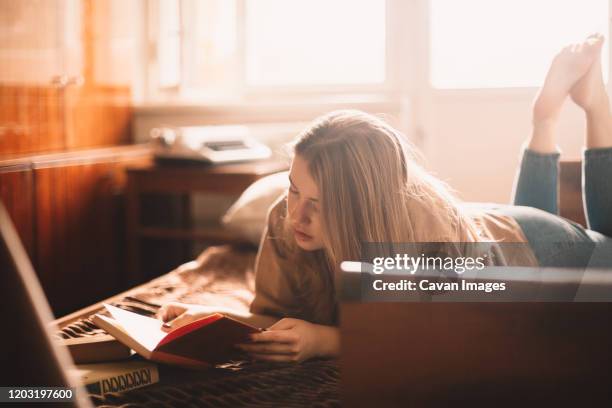 young woman reading book while lying on bed at home - allongé sur le devant photos et images de collection