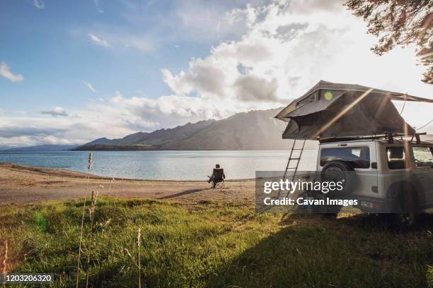 a woman sits along a lake next to a suv with a tent in new zealand - suv berg stock-fotos und bilder