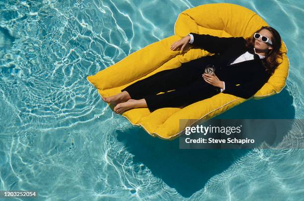 Portrait of American actress Diane Lane, barefoot and dressed in a suit and bowtie & sunglasses, as she floats in a pool on an inflatable chair, Los...