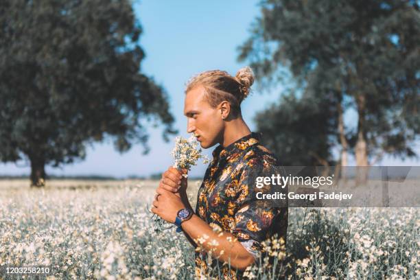 portrait of curly blond guy is standing at the field and sniffs a flowers. romantic, summer time. flowers around and trees. - tee mockup stock pictures, royalty-free photos & images