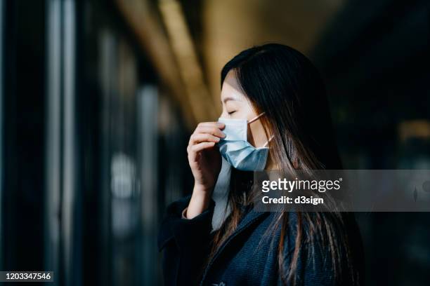 asian woman with eyes closed wearing protective face mask commuting in the city and waiting for subway in platform - woman cough stock-fotos und bilder