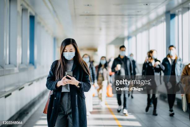 asian woman with protective face mask using smartphone while commuting in the urban bridge in city against crowd of people - epidemie stock-fotos und bilder