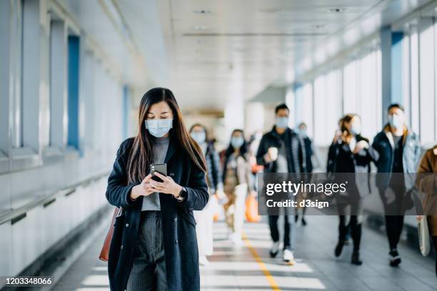asian woman with protective face mask using smartphone while commuting in the urban bridge in city against crowd of people - masque de chirurgien photos et images de collection