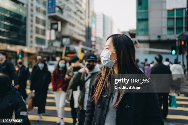 asian woman with protective face mask crossing road in busy downtown city street against crowd of pedestrians and highrise urban buildings - epidemic ストックフォトと画像