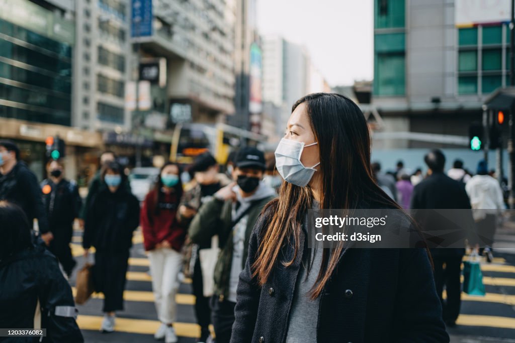 Asian woman with protective face mask crossing road in busy downtown city street against crowd of pedestrians and highrise urban buildings