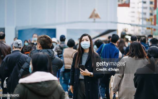 asian woman with protective face mask holding smartphone standing in the middle of busy downtown city street amidst crowd of pedestrians passing by - crowded stock pictures, royalty-free photos & images