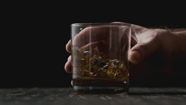 Glass with whiskey rotating at wooden table. Close-up of whiskey with ice.