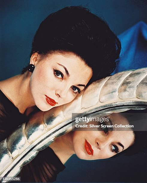 Joan Collins, British actress, posing beside a mirror relects an image of her face, in a studio portrait, against a blue background, circa 1955.