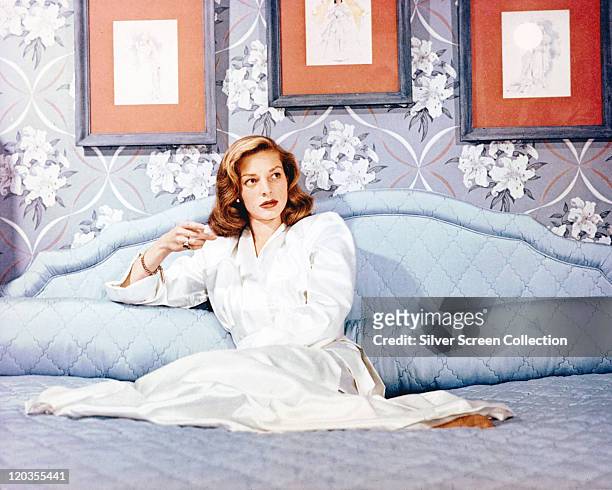 Lauren Bacall, US actress, wearing a long white dressing gown while reclining on a bed, with three pictures hanging on the wall behind her, circa...