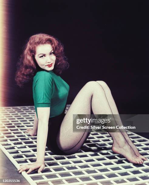 Julie Newmar, US actress, dancer and singer, sitting on a white mat, wearing a green short-sleeved, woollen top and fishnet stockings, circa 1955.