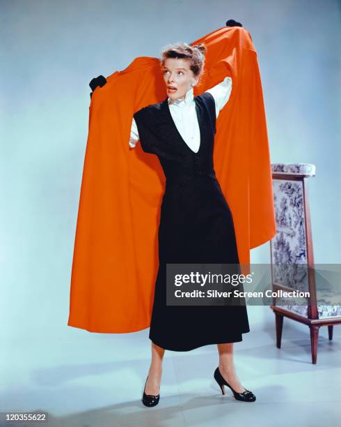 Katharine Hepburn , US actress, wearing a black dress as she puts on a red coat in a studio portrait, against a white background, 1965.