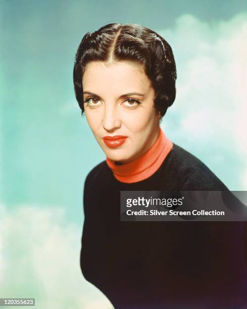 Katy Jurado , Mexican actress, wearing red polo neck top beneath a black jumper in a studio portrait, against a background of blue sky and clouds,...