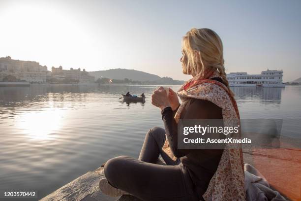 reife frau sitzt auf einem ghat bei sonnenaufgang mit einem heißen getränk, blickt über den see - blonde attraction stock-fotos und bilder