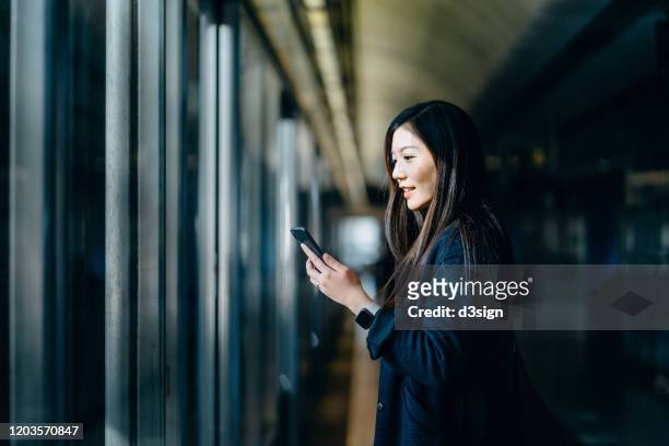 asian woman reading on smartphone while commuting in the city and waiting for subway in platform - andén de estación de metro fotografías e imágenes de stock