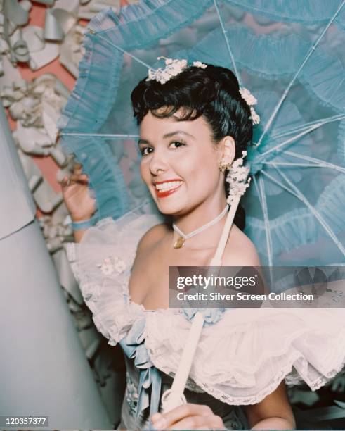 Lena Horne , US jazz singer and actress, wearing an off-the-shoulder dress with a white lace ruff over the shoulders, smiling while carrying a blue...