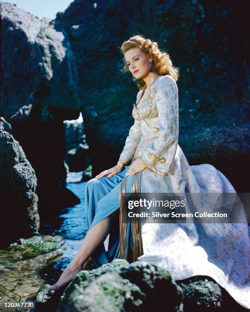 Maureen O'Hara, Irish actress, wearing a pale blue, gold brocade jacket, with a blue skirt, sitting barefoot on a rock, circa 1950.