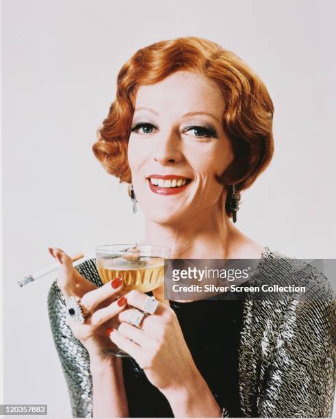 Maggie Smith, British actress, smiling in a studio portrait, weaing a black top beneath a silver-and-black cardigan, holding a lit cigarette and a...