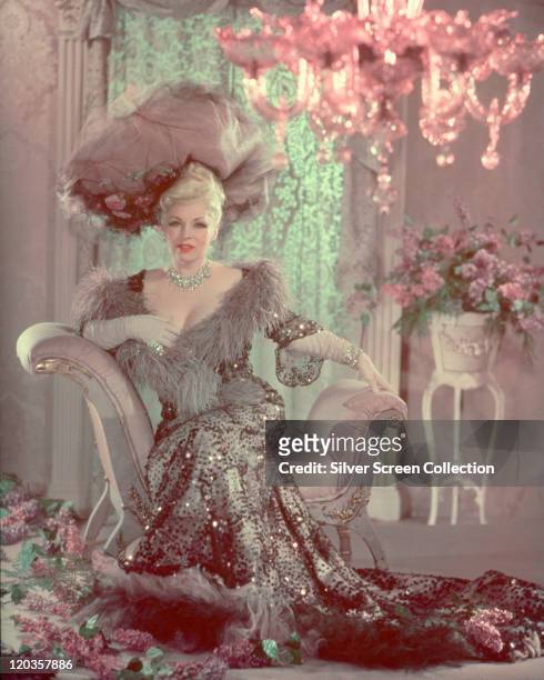 Mae West , US actress, in period costume , sitting on a backless armchair in a studio portrait, with pink flowers in the background and strewn across...