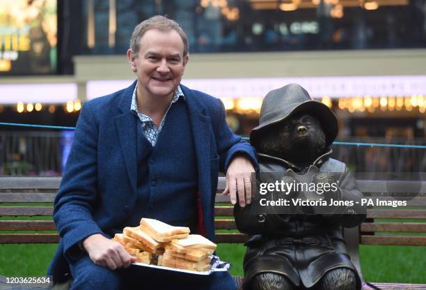 Hugh Bonneville eating a marmalade sandwich alongside a Paddington Bear statue as he attends the Scenes In The Square statue trail unveiling in...