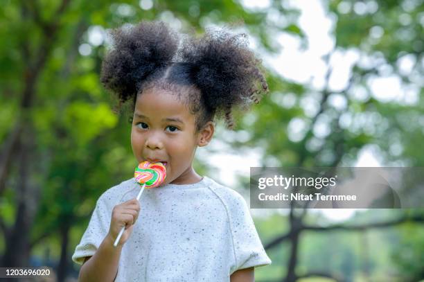 cute little afro america girl eating lollipop in the park. - lolly stockfoto's en -beelden