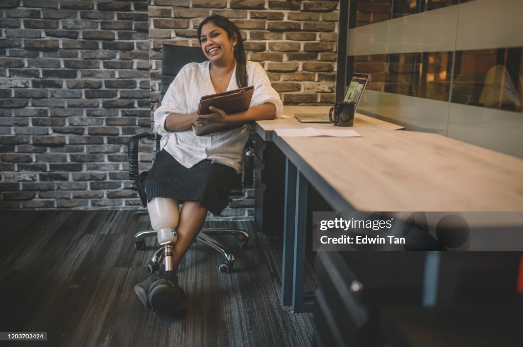 A portrait of an asian indian white collar worker with prosthetic leg sitting at her workstation with toothy smile confidence holding her digital tablet with leg crossed on ankle