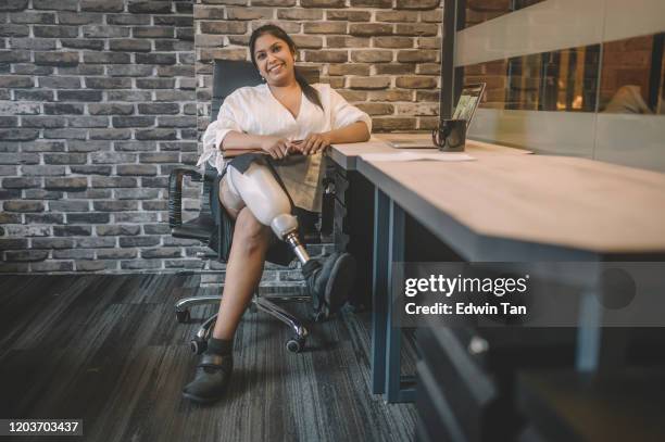 a portrait of an asian indian white collar worker with prosthetic leg sitting at her workstation with toothy smile confidence holding her digital tablet with leg crossed on knee - injured woman stock pictures, royalty-free photos & images