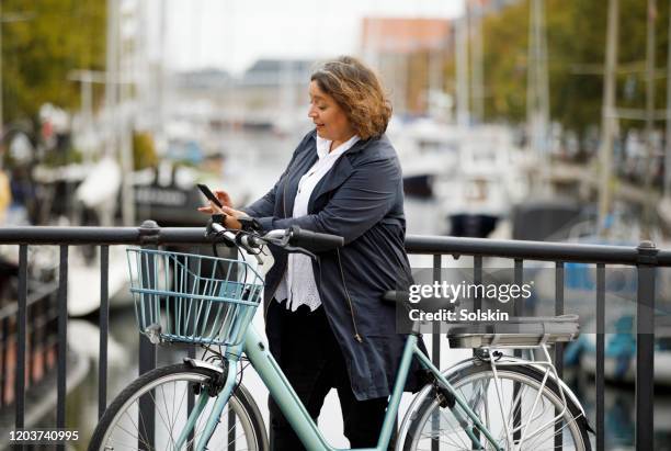 woman standing by electric bicycle in city area looking at smartphone - ebike stockfoto's en -beelden