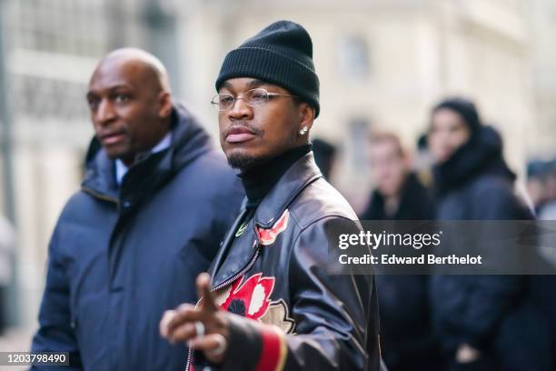 Singer Ne-Yo wears a black wool beanie hat, earrings, glasses, a necklace, a black turtleneck pullover, a black and red leather jacket with printed...