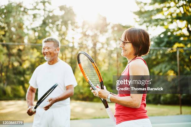 laughing senior friends in discussion at net during early morning tennis match - anziani attivi foto e immagini stock