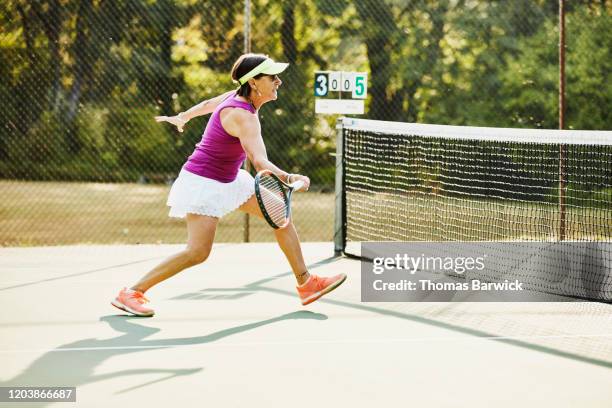 senior woman preparing to hit shot at net during early morning tennis match - sun visor stock pictures, royalty-free photos & images