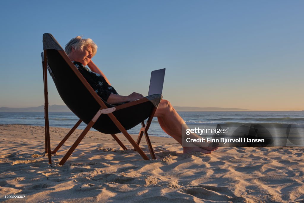 Senior woman sitting in a chair at the beach at sunset working on her computer