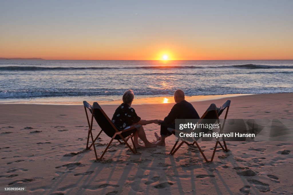 Senior couple sitting on the beach hand in hand watching the sunset over the atlantic ocean