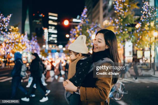 young mother with little daughter walking in downtown city street at night, against illuminated and multi-coloured street lights and urban buildings - asien metropole nachtleben stock-fotos und bilder