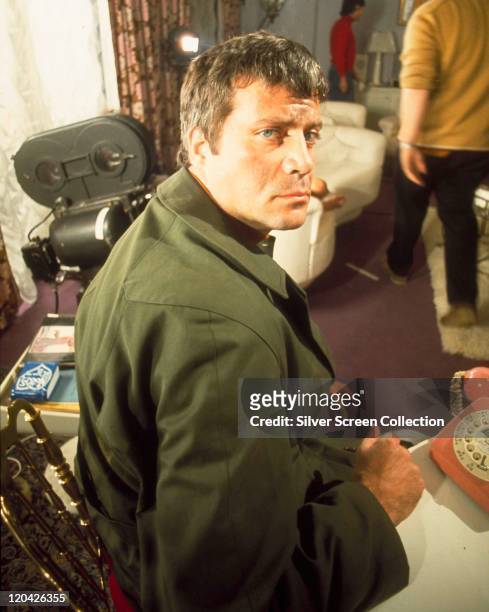 Oliver Reed , looking over his shoulder, wearing a green coat on the set of a film, with a movie camera in the backround, circa 1970.