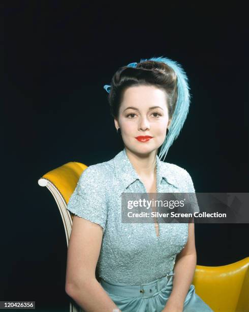 Olivia de Havilland, British actress, wearing a blue dress with a matching blue feather in her hair, sitting on a yellow sofa in a studio portrait,...