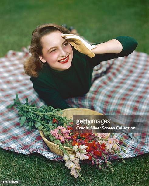 Olivia de Havilland, British actress, wearing a green jumper and gardening gloves she lays on a tartan rug with a basket of snapdragons, circa 1945.