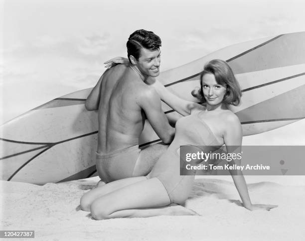 young couple with surfboard on beach, smiling - 60s man stock pictures, royalty-free photos & images