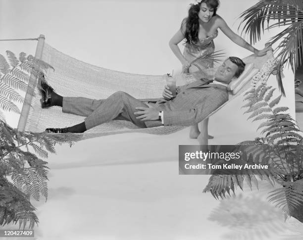 young woman standing beside man relaxing in hammock with drink - 1962 fotografías e imágenes de stock