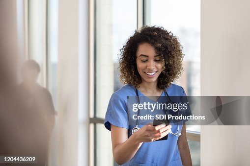 Fröhliche Krankenschwester überprüft Nachrichten auf dem Smartphone, während im Krankenhaus Flur Fröhliche Krankenschwester überprüft Nachrichten auf dem Smartphone, während im Krankenhaus Flur