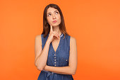 Pensive brunette woman in denim dress looking up with thoughtful doubtful expression